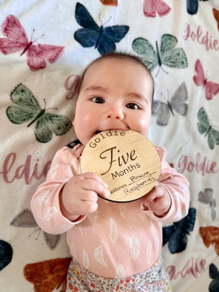Baby holding a 'Five Months' milestone wooden plaque on a butterfly-patterned blanket.