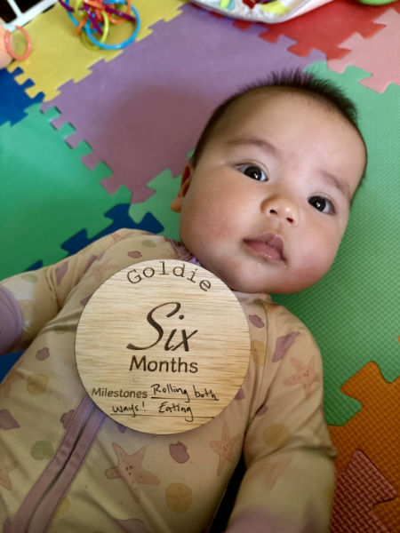 Baby lying on a colorful play mat with a wooden milestone card reading 'Goldie Six Months'.
