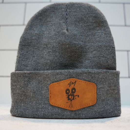 A dark gray heather knit beanie hat with a folded cuff, featuring a brown faux-leather patch on the front with a black graphic logo and handwritten text.