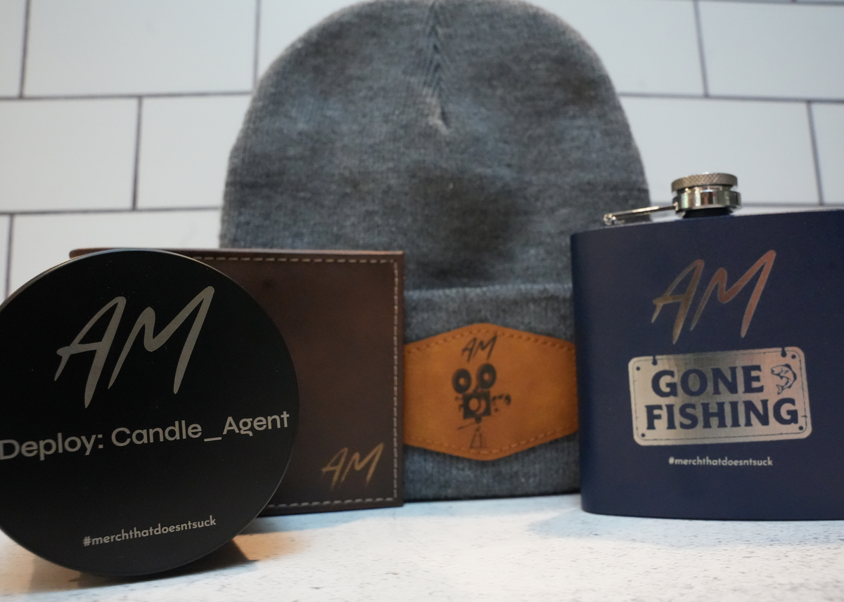 A curated gift set featuring a gray knit beanie with a brown leather patch, a navy blue flask with a silver cap, a black round tin, and a brown leather wallet, all branded with 'AM' and 'Deploy: Candle_Agent' on a white tiled background.