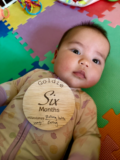 Baby lying on a colorful play mat with a wooden milestone card reading 'Goldie Six Months'.